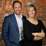 Beth and Ryan Waller, Guelph real estate agents, smiling together against a rustic brick wall backdrop, representing their expertise and approachability in the Guelph real estate market.
