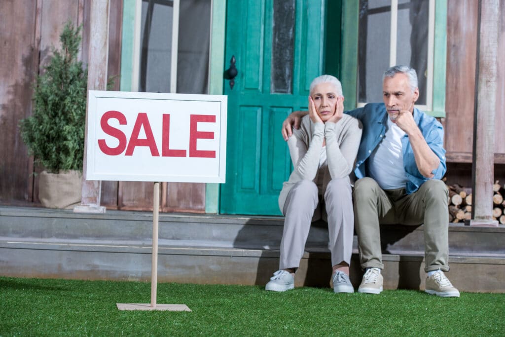 Couple sitting on steps with worried expressions beside a "SALE" sign, illustrating the emotional challenges of selling a home in Guelph.